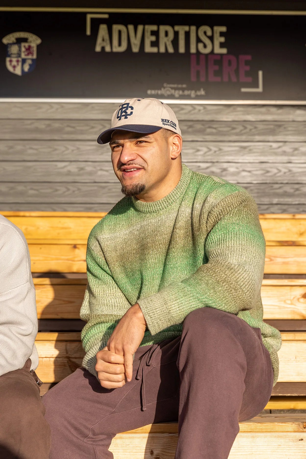 Rex Club | A man sits on wooden bleachers in sunlight, wearing a green 100% cotton sweater, brown pants, and a beige Rex Club - TwoTone Canvas Cap by Rex Club. A sign behind him reads "ADVERTISE HERE. | Custom Caps | Custom Hats | Team Headwear | UK