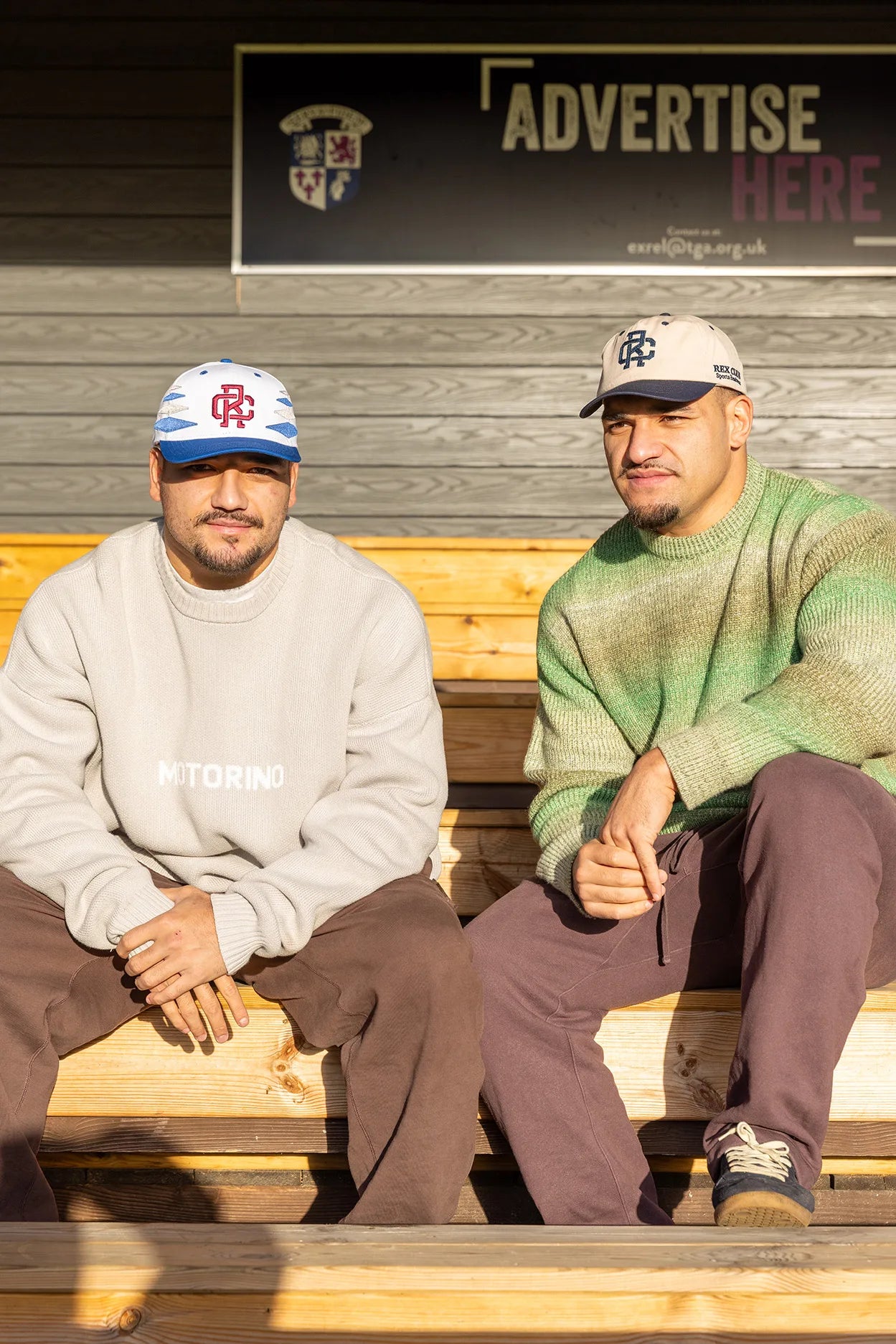 Rex Club | Two men sit on wooden benches in sunlight, each wearing Rex Club - TwoTone - Canvas Caps and sweaters with brown pants. A wall with a partially visible “ADVERTISE HERE” sign is behind them. | Custom Caps | Custom Hats | Team Headwear | UK