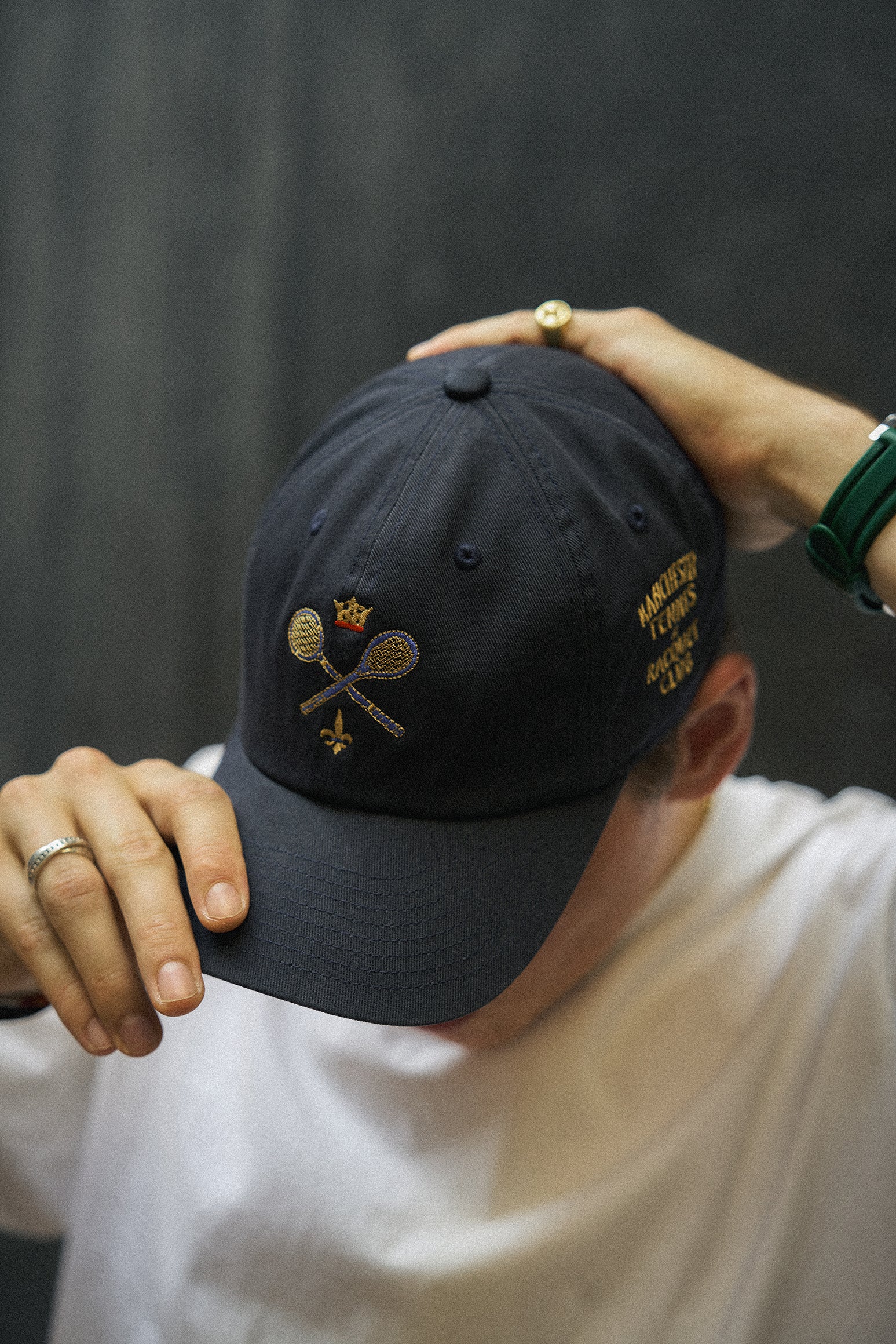 Rex Club | A person wearing the Rex Club MTRC - Navy cap—an iconic Manchester Tennis & Racquets Club piece made from 100% cotton and featuring embroidered tennis rackets, a crown, and text—is shown holding the cap’s brim and top while dressed in a white shirt. | Custom Caps | Custom Hats | Team Headwear | UK