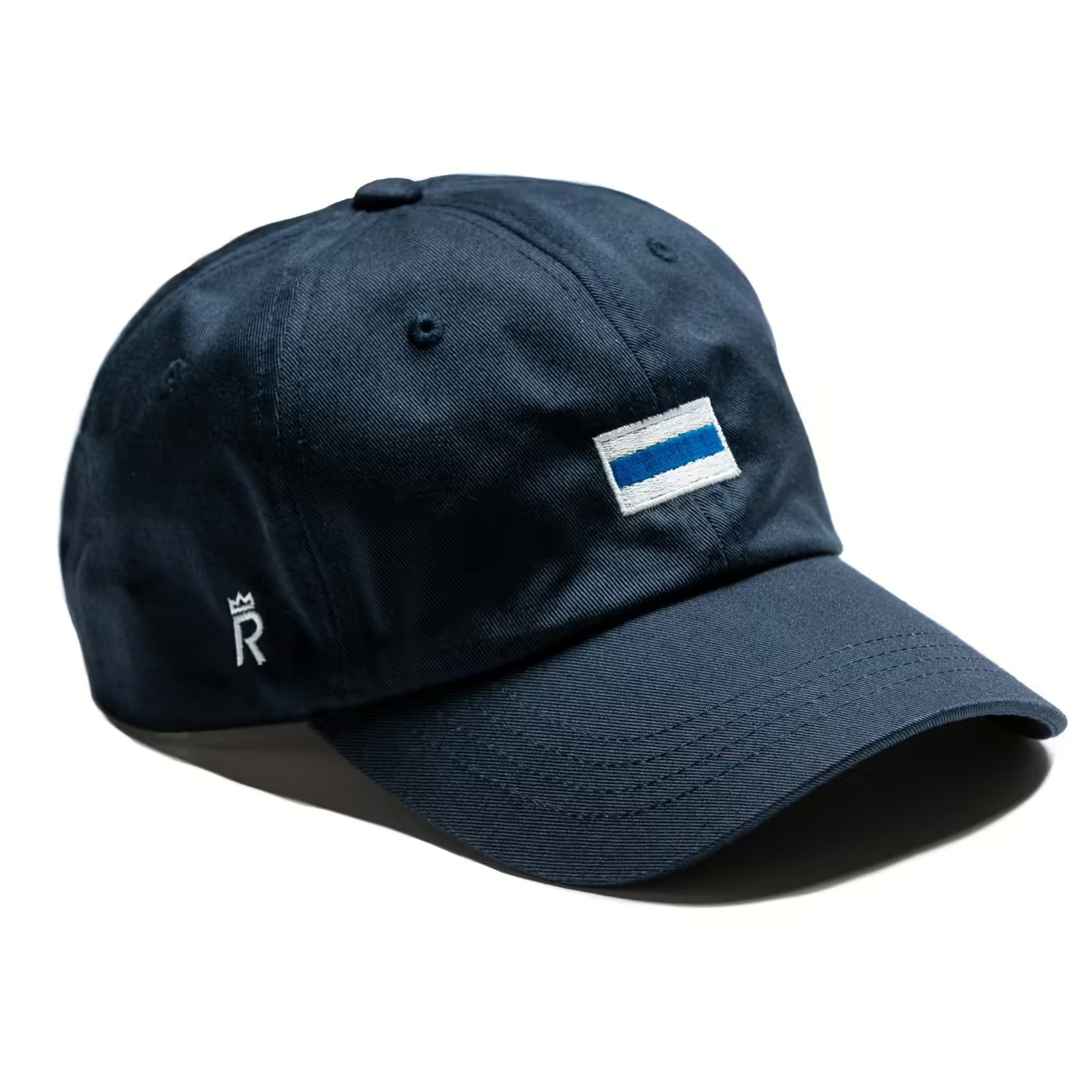 Rex Club | The Club Colours slouch cap by Rex Club is navy blue with a small rectangular patch of blue and white stripes on the front, classic club colours, plus a white crown and “R” logo on the left side. | Custom Caps | Custom Hats | Team Headwear | UK