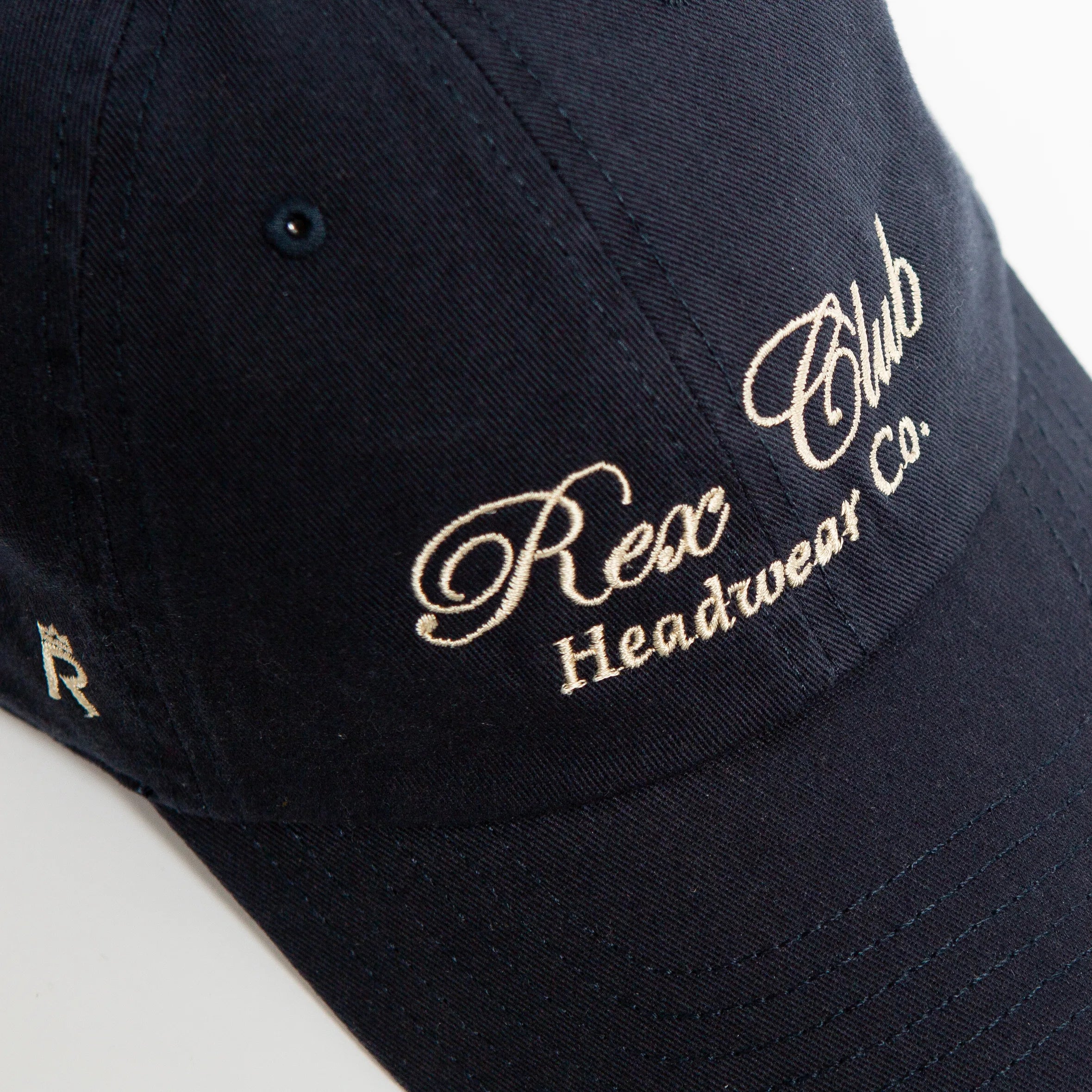 Rex Club | A navy blue 100% cotton baseball cap from Rex Club, featuring "Rex Club Headwear Co." embroidered in cream cursive on the front, displayed at an angle against a white background. | Custom Caps | Custom Hats | Team Headwear | UK