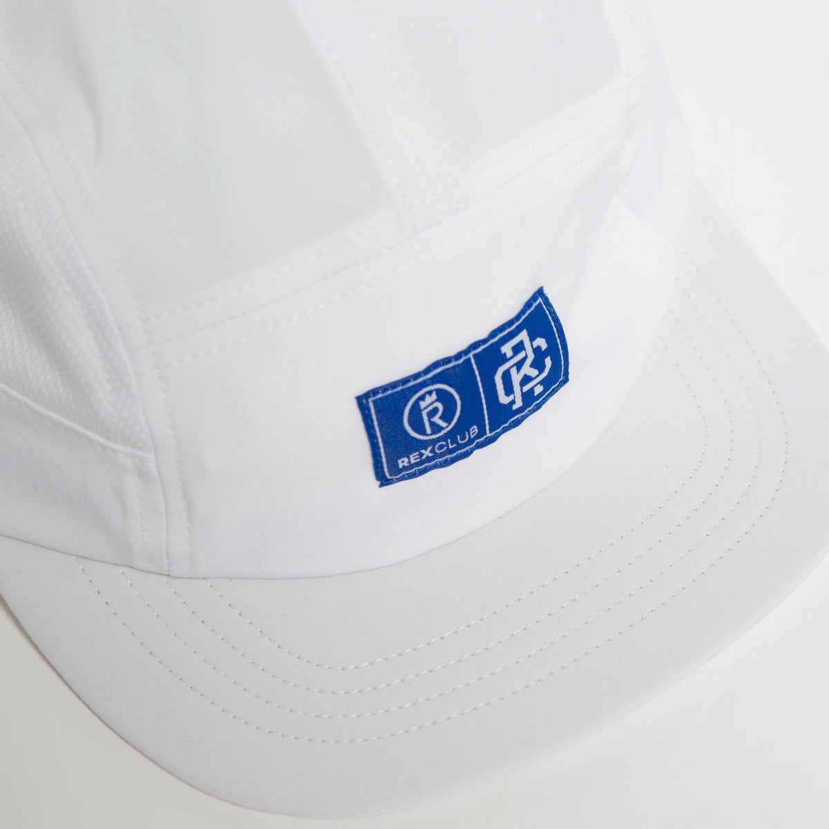 Rex Club | A close-up of a Rex Club Running Cap in white polyester, featuring a blue patch with "REXCLUB" and two white logos—an "R" in a circle and a geometric design. Finished with subtle black accents. | Custom Caps | Custom Hats | Team Headwear | UK