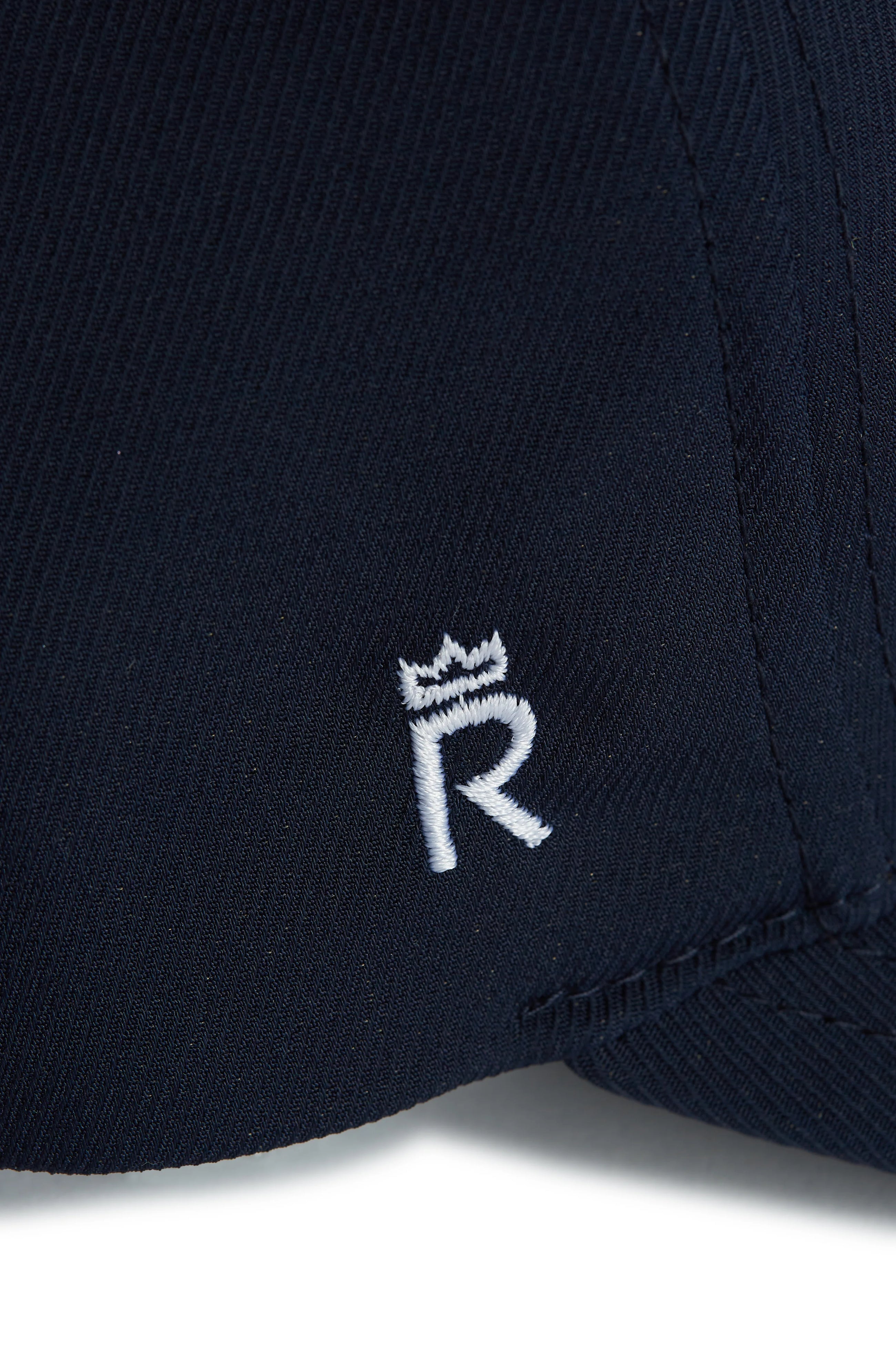 Rex Club | Close-up of dark navy fabric from the Rex Club OFFSEASON 26 - Navy performance cap, featuring a white embroidered letter "R" topped by a small crown symbol. The stitching and fabric texture are clearly visible, emphasizing the premium quality of this signature Rex Club design. | Custom Caps | Custom Hats | Team Headwear | UK