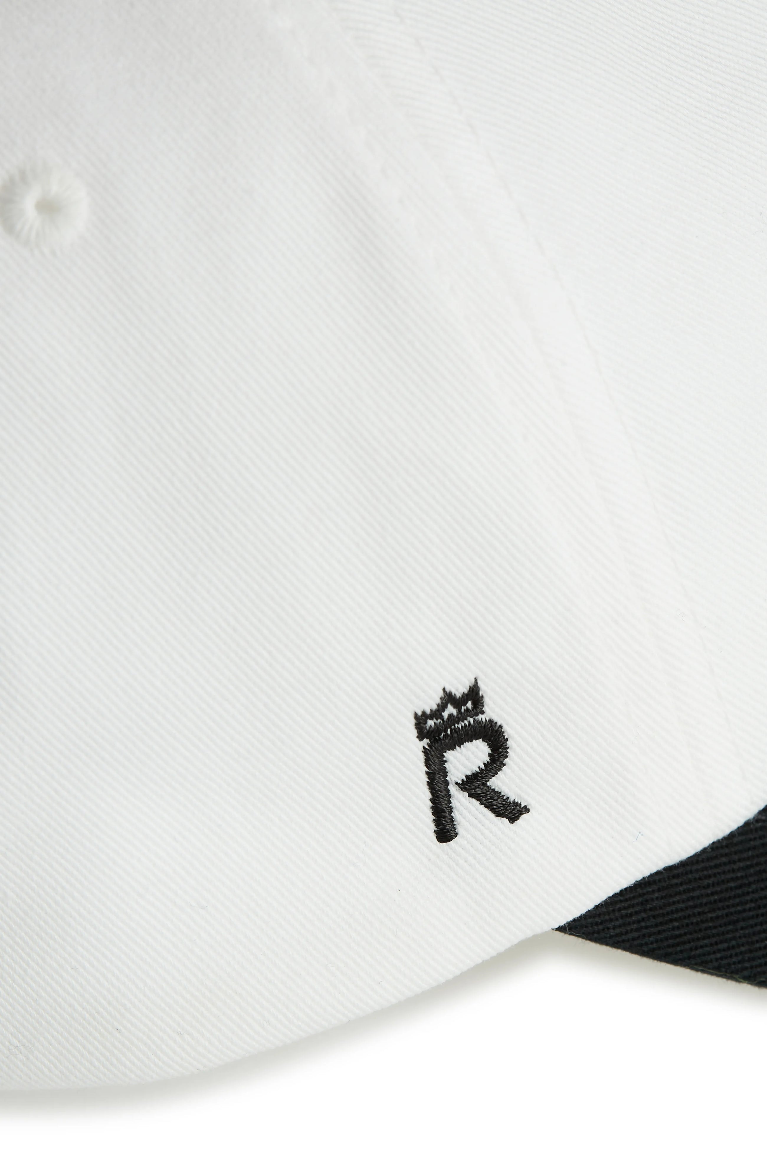 Rex Club | Close-up of Rex Club's Goat - Black & White cap in white fabric, showcasing a small black embroidered "R" crowned near the brim, with part of the black underside also visible. | Custom Caps | Custom Hats | Team Headwear | UK
