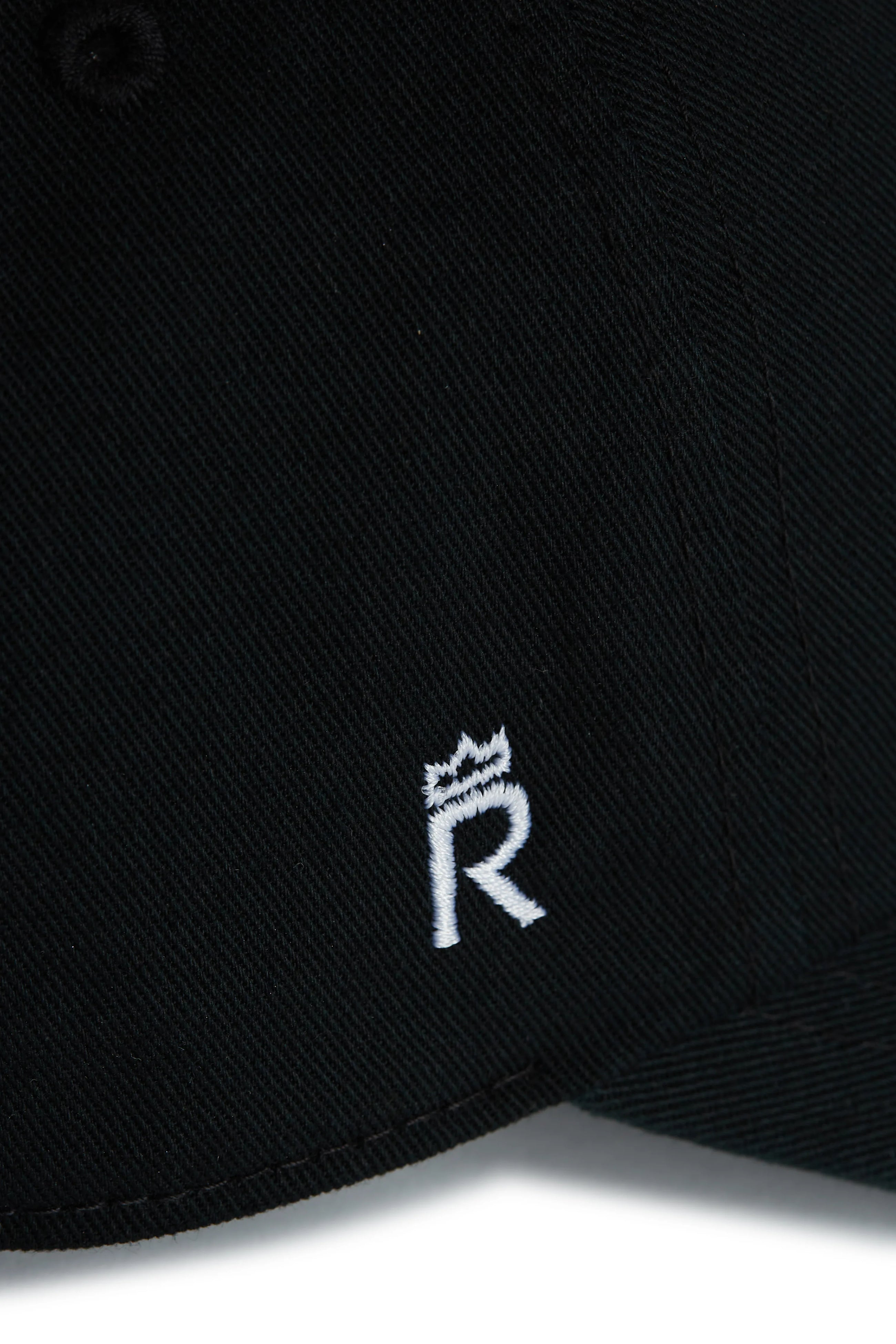 Rex Club | Close-up of the Goat - Black cap by Rex Club, showcasing a small white embroidered letter "R" with a crown accent on black fabric. | Custom Caps | Custom Hats | Team Headwear | UK