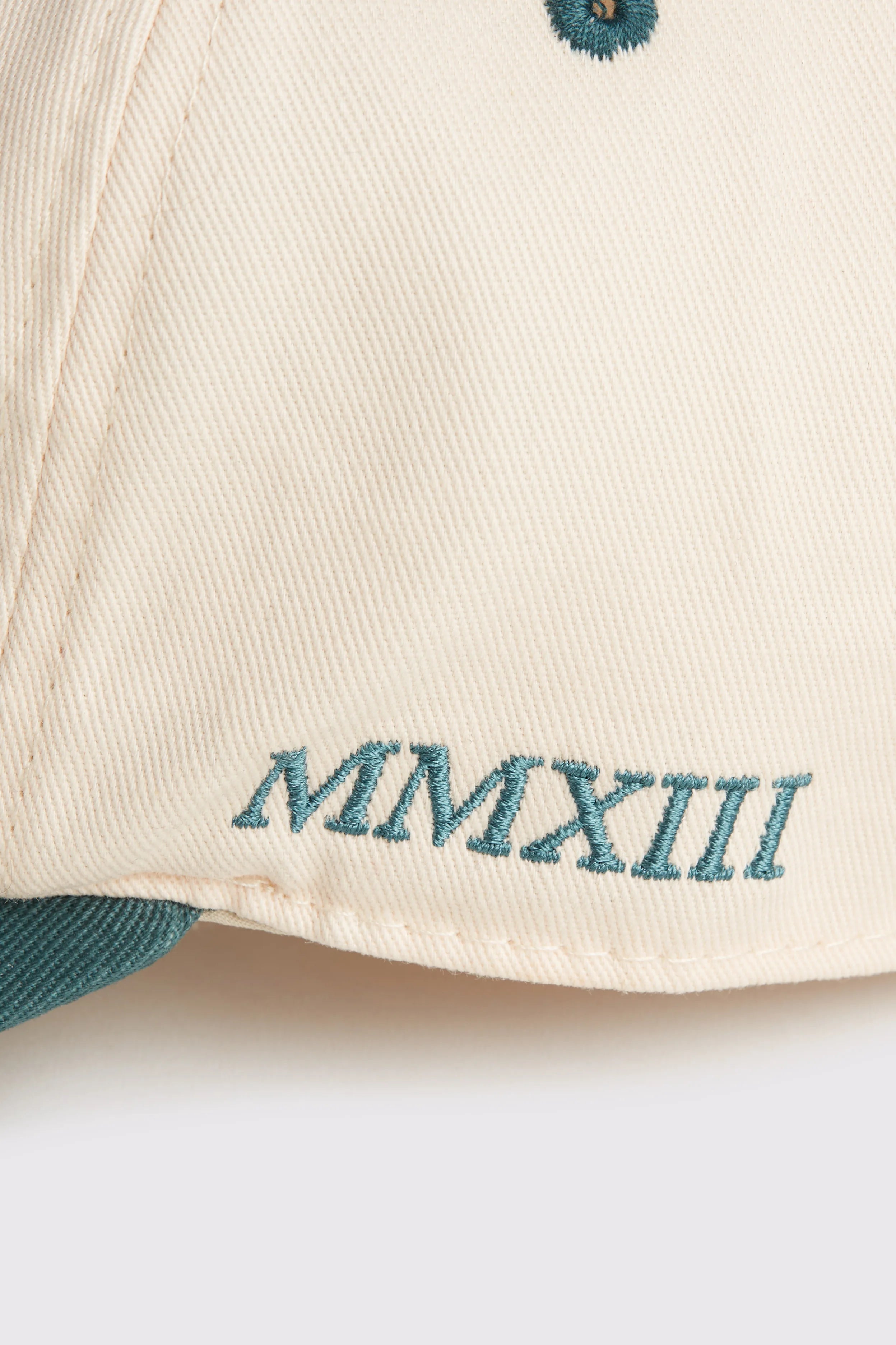 Rex Club | A close-up of the RC - Two/Tone - SS26 Green cap by Rex Club, crafted from 100% cotton in beige, featuring "MMXIII" embroidered in teal thread near the brim. | Custom Caps | Custom Hats | Team Headwear | UK