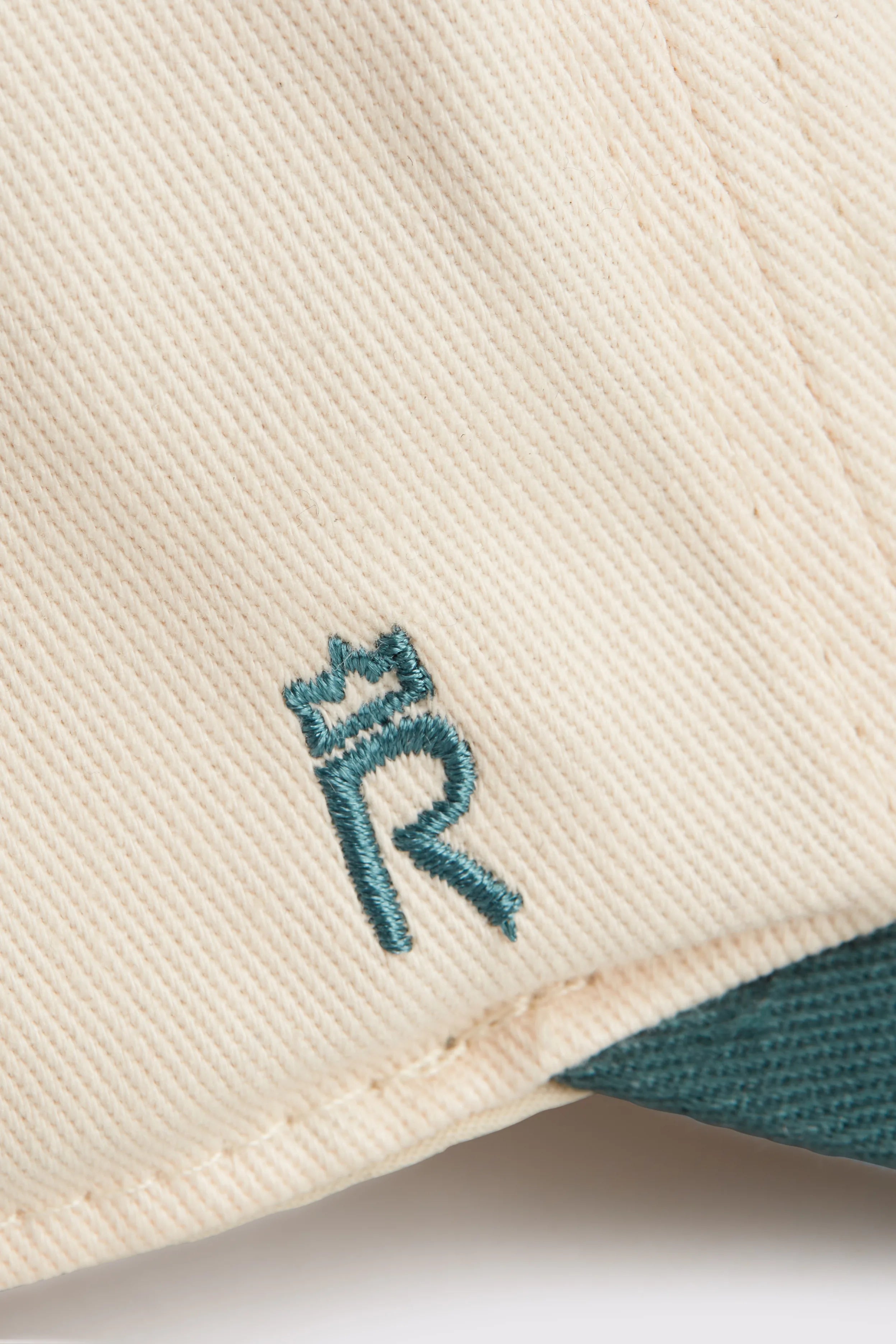 Rex Club | Close-up of RC - Two/Tone - SS26 Green by Rex Club: cream-colored 100% cotton fabric with a teal embroidered "R" topped by a crown, and a teal edge in the lower right corner. | Custom Caps | Custom Hats | Team Headwear | UK