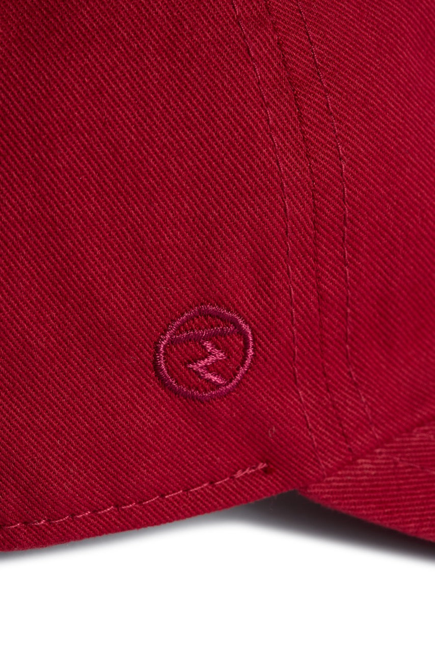 Rex Club | Close-up of the Rex Club “In the details - Tyrian” baseball cap in red 100% cotton, showcasing detailed stitching and a small embroidered circle with a zigzag or “Z” shape inside it. | Custom Caps | Custom Hats | Team Headwear | UK