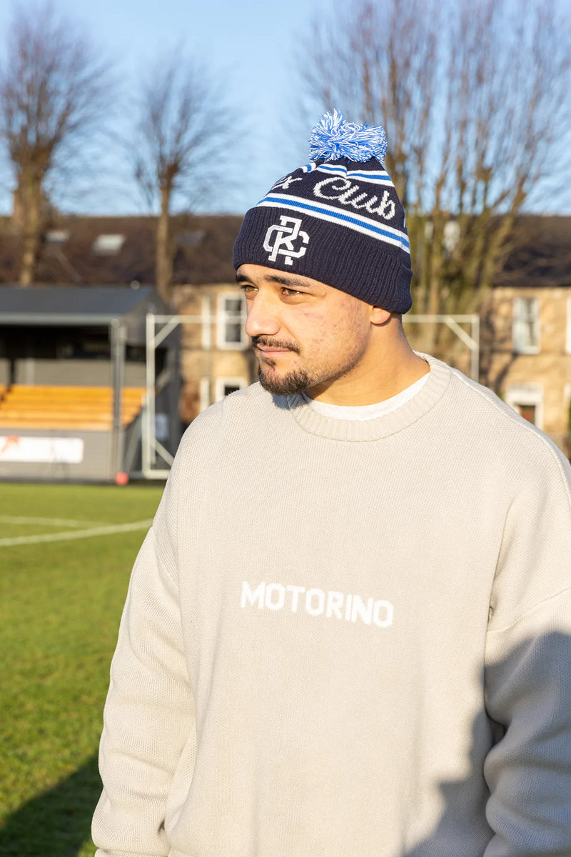 Rex Club | A man wearing a blue Rex Club Bobble Hat and a light gray "MOTORINO" sweatshirt stands on a grassy sports field in sunlight, with buildings and leafless trees in the background. | Custom Caps | Custom Hats | Team Headwear | UK