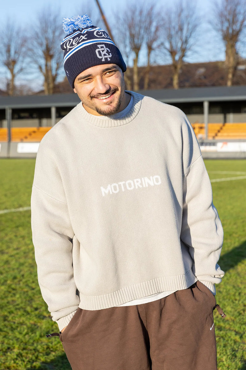 Rex Club | A smiling man wears a Rex Club Bobble Hat and a light beige sweater with "MOTORINO" text, paired with brown pants, standing on a grassy sports field with bleachers and leafless trees in the background. | Custom Caps | Custom Hats | Team Headwear | UK