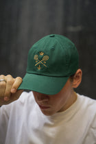 Rex Club | A person wearing the Rex Club MTRC - Green cap, featuring embroidery of two crossed tennis rackets, a crown, and the year “1876,” looks down while holding the brim. They are dressed in a white shirt against a dark, blurred background. | Custom Caps | Custom Hats | Team Headwear | UK