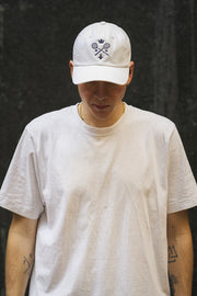 Rex Club | A person wearing the Rex Club MTRC - White 100% cotton T-shirt and a matching white baseball cap with black symbols, reminiscent of Manchester Tennis & Racquets Club caps, stands against a dark background, looking down. Tattoos are visible on both arms. | Custom Caps | Custom Hats | Team Headwear | UK