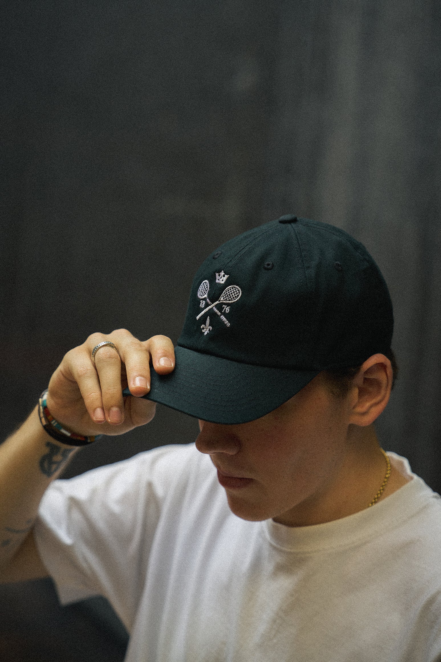 Rex Club | A person wearing the Rex Club MTRC - Black cap, featuring tennis racquets and a crown logo, paired with a white t-shirt and a gold necklace, is adjusting the visor. The dark, blurred background highlights the stylish Rex Club tennis merchandise. | Custom Caps | Custom Hats | Team Headwear | UK