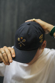 Rex Club | A person wearing the Rex Club MTRC - Navy cap—an iconic Manchester Tennis & Racquets Club piece made from 100% cotton and featuring embroidered tennis rackets, a crown, and text—is shown holding the cap’s brim and top while dressed in a white shirt. | Custom Caps | Custom Hats | Team Headwear | UK