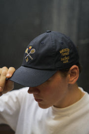 Rex Club | A person in a white shirt holds the brim of the Rex Club MTRC - Navy cap, a navy blue hat embroidered with crossed tennis racquets and “Manchester Tennis Racquet Club” text. Crafted from 100% cotton, these caps combine comfort and style. | Custom Caps | Custom Hats | Team Headwear | UK
