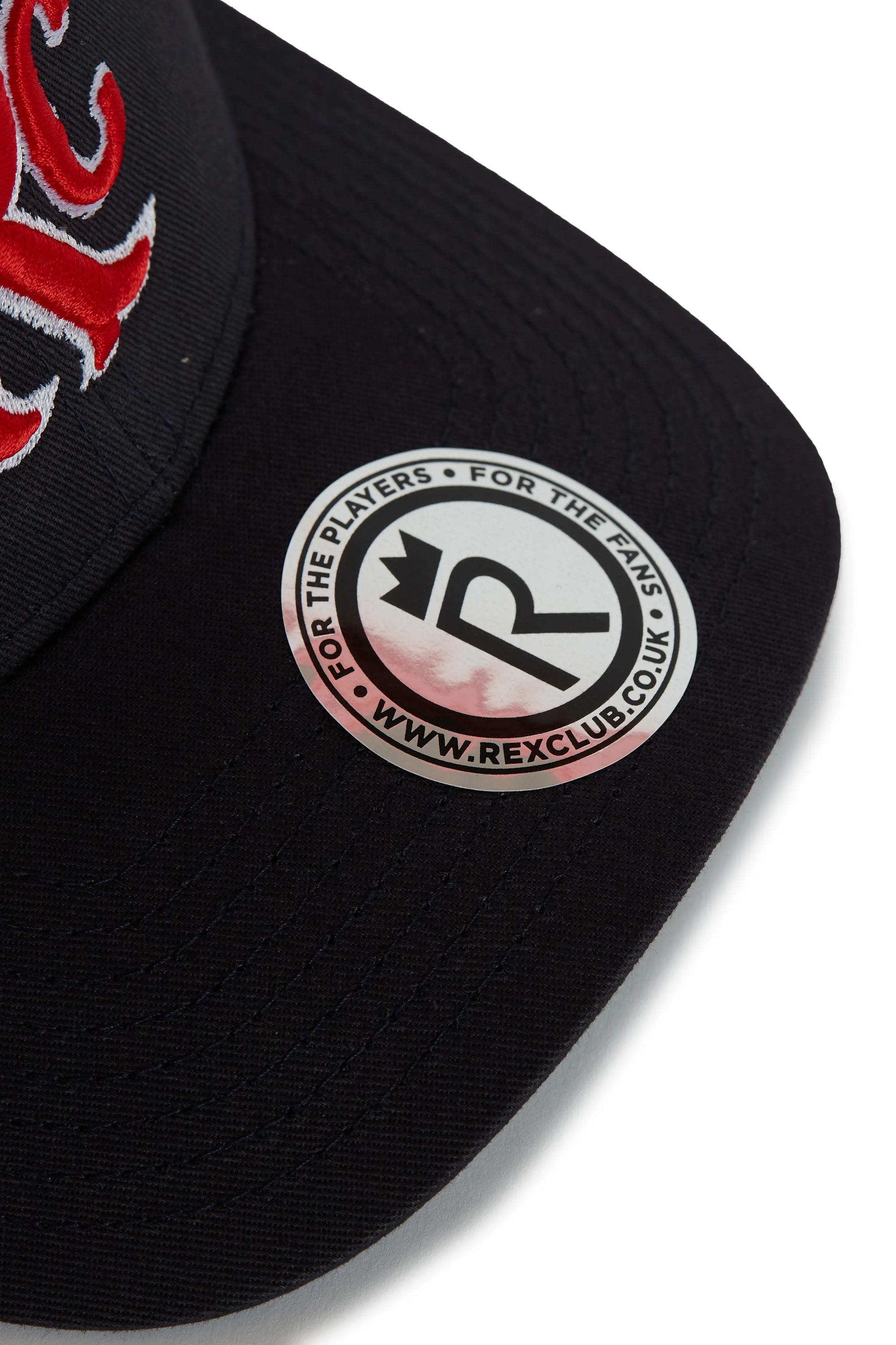 Rex Club | A close-up of the black RC 26 Trucker hat by Rex Club, featuring red embroidery, a sticker on the brim with “FOR THE PLAYERS • FOR THE FANS” and “WWW.REXCLUB.COM,” and a sport team "R" monogram with an arrow. | Custom Caps | Custom Hats | Team Headwear | UK