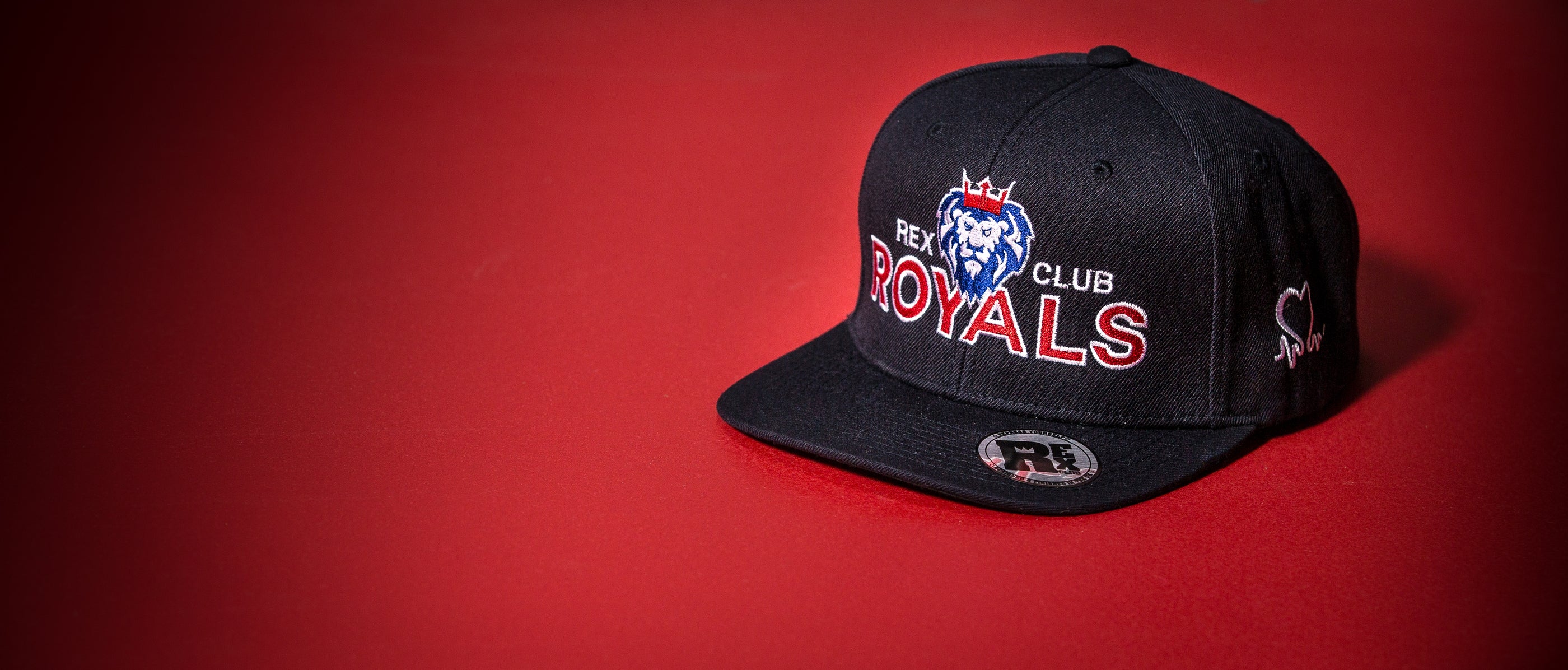 Rex Club | A black baseball cap with a blue lion logo and the words "REXY CLUB ROYALS" embroidered on the front, displayed on a red surface. | Custom Caps | Custom Hats | Team Headwear | UK
