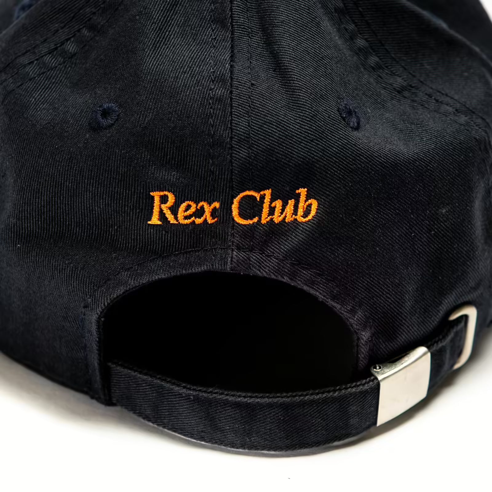 Rex Club | A close-up of the back of a black OFFSEASON 23 Slouch baseball cap by Rex Club, made from 100% cotton, with "Rex Club" embroidered in orange above the adjustable strap and silver buckle. | Custom Caps | Custom Hats | Team Headwear | UK