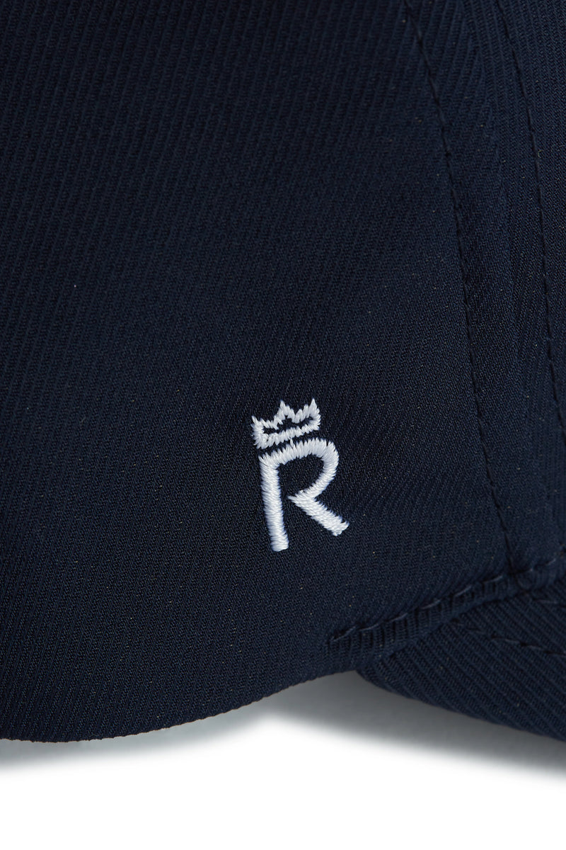 Rex Club | Close-up of dark navy fabric from the Rex Club OFFSEASON 26 - Navy performance cap, featuring a white embroidered letter "R" topped by a small crown symbol. The stitching and fabric texture are clearly visible, emphasizing the premium quality of this signature Rex Club design. | Custom Caps | Custom Hats | Team Headwear | UK