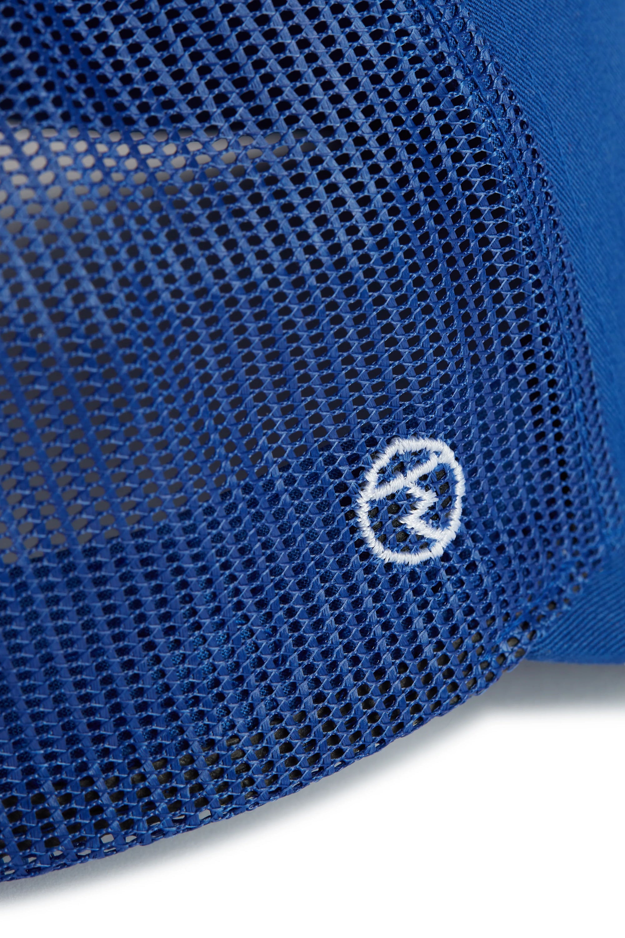Rex Club | Close-up of blue mesh fabric from the RC 26 Trucker by Rex Club, showcasing a small white circular embroidered logo with a stylized lightning bolt at the center. | Custom Caps | Custom Hats | Team Headwear | UK