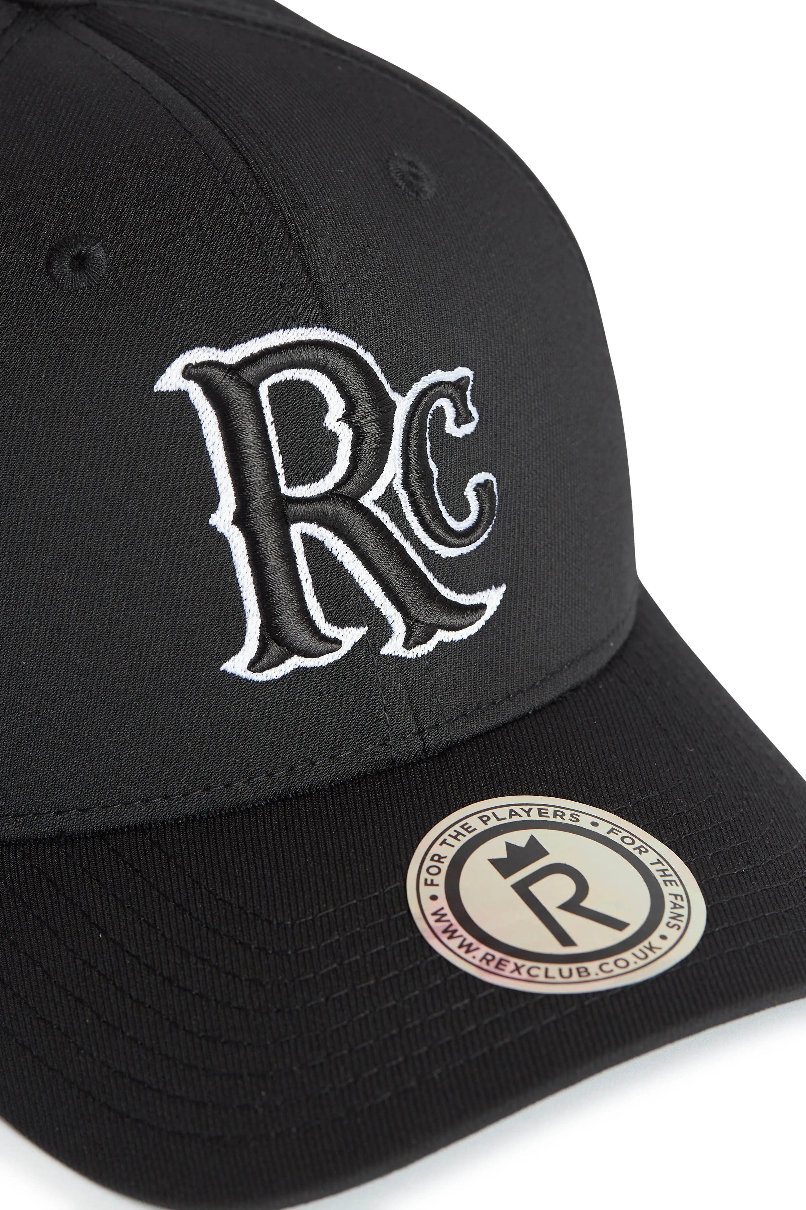 Rex Club | A close-up of the RC 26 Performance black baseball cap by Rex Club, featuring a large white "RC" monogram on the front and a round brim sticker reading "FOR THE PLAYERS • FOR THE REAL" and "www.REXCLUB.CO.UK. | Custom Caps | Custom Hats | Team Headwear | UK