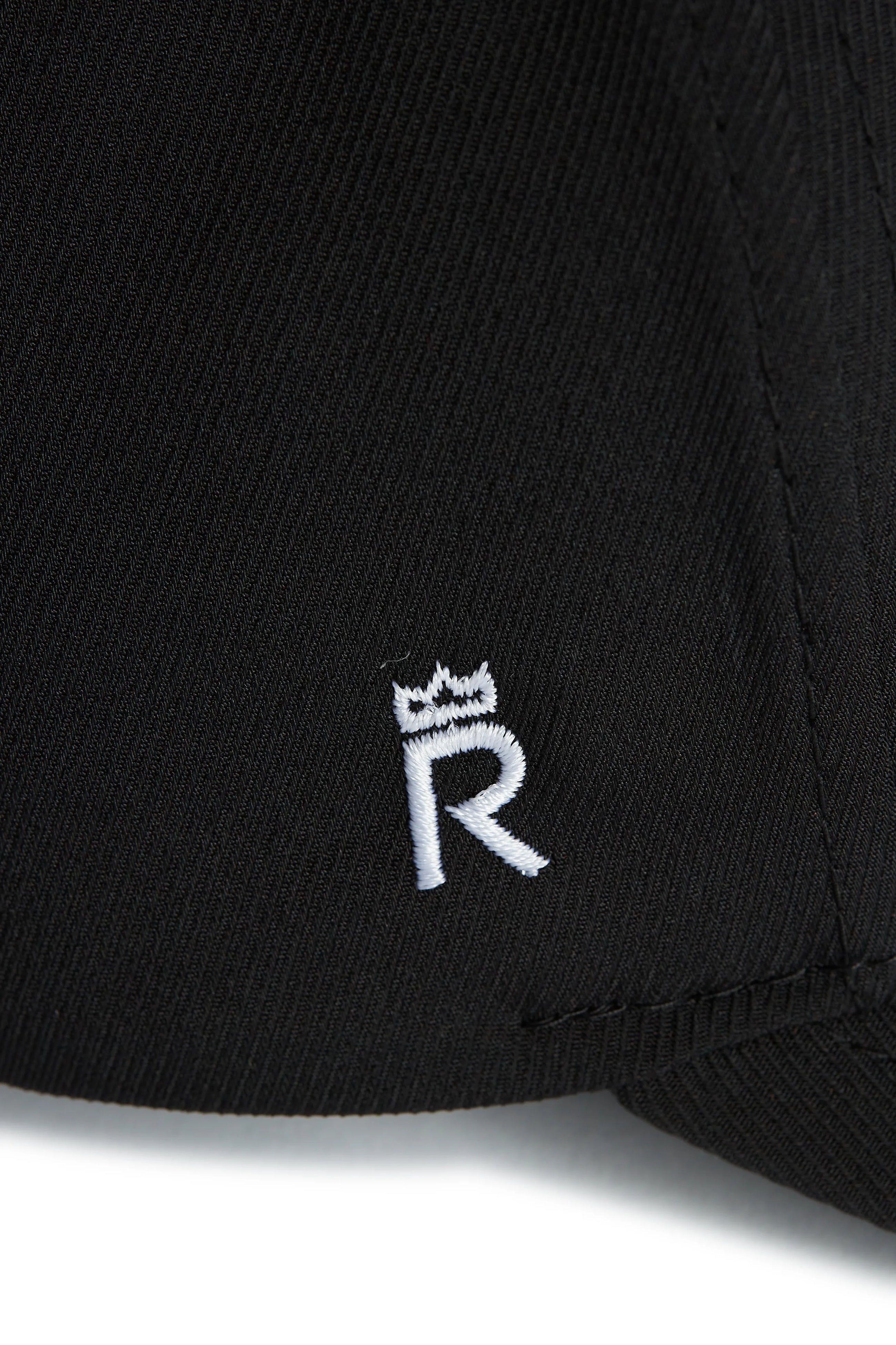 Rex Club | Close-up of RC 26 Performance black jersey by Rex Club, highlighting a white embroidered “R” monogram crowned with a small symbol—signature detail on sports apparel. | Custom Caps | Custom Hats | Team Headwear | UK