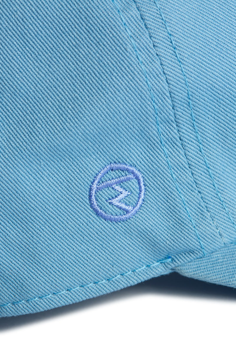 Rex Club | Close-up of light blue fabric from the Rex Club “In the details – Cornflower” collection, featuring a stitched circular emblem with a stylized ‘W’ or zigzag shape at its center. The stitching and lightweight, breathable material suggest it is part of cotton caps or other summer hats. | Custom Caps | Custom Hats | Team Headwear | UK