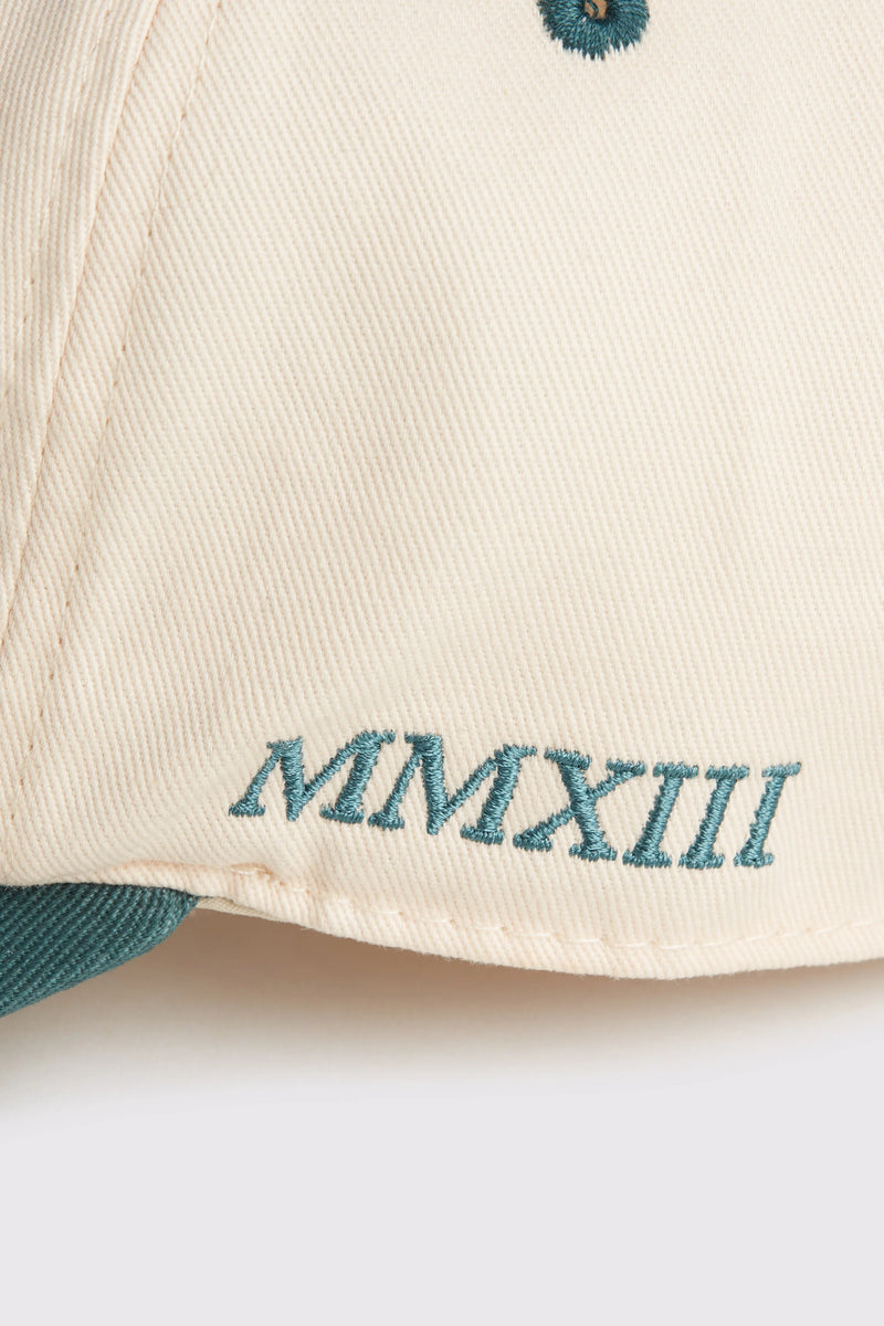 Rex Club | A close-up of the RC - Two/Tone - SS26 Green cap by Rex Club, crafted from 100% cotton in beige, featuring "MMXIII" embroidered in teal thread near the brim. | Custom Caps | Custom Hats | Team Headwear | UK