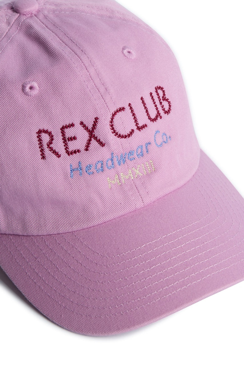 Rex Club | Introducing the "In the details - Parma Violet" baseball cap by Rex Club. This slouch cap comes in a light pink shade, crafted from 100% cotton, and features "REX CLUB Headwear Co. PARIS" embroidered across the front in vibrant red, blue, and yellow threads. Perfect for summer days, it is shown here resting on a white surface. | Custom Caps | Custom Hats | Team Headwear | UK