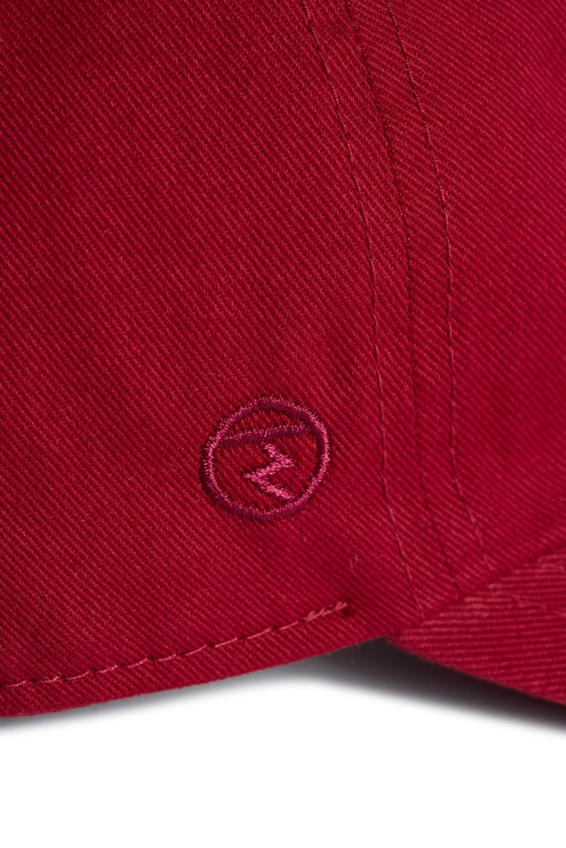 Rex Club | Close-up of the Rex Club “In the details - Tyrian” baseball cap in red 100% cotton, showcasing detailed stitching and a small embroidered circle with a zigzag or “Z” shape inside it. | Custom Caps | Custom Hats | Team Headwear | UK