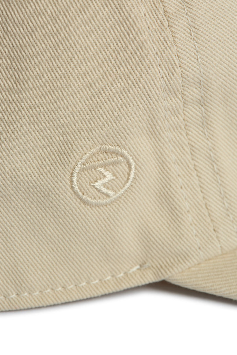 Rex Club | Close-up of beige 100% cotton fabric from Rex Club’s “In the details - Cotton” collection, featuring visible stitching and a small embroidered logo—a circle with a zigzag line inside—commonly seen on their stylish slouch caps and summer hats. | Custom Caps | Custom Hats | Team Headwear | UK